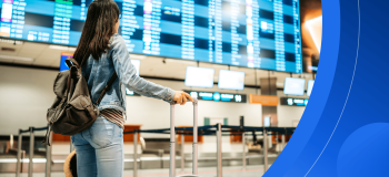 A female international student wearing casual clothes and holding luggage stands in front of a large screen with flight information at an airport.